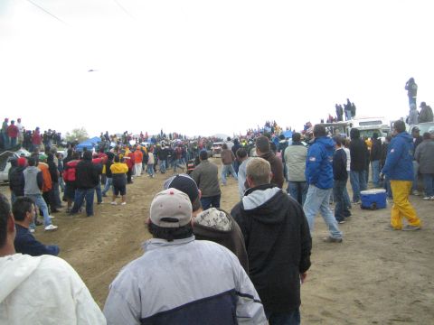 1990 Ford Bronco - Spectators in cold-weather gear line a sandy off-road course, with blue tents and trucks visible in the background.