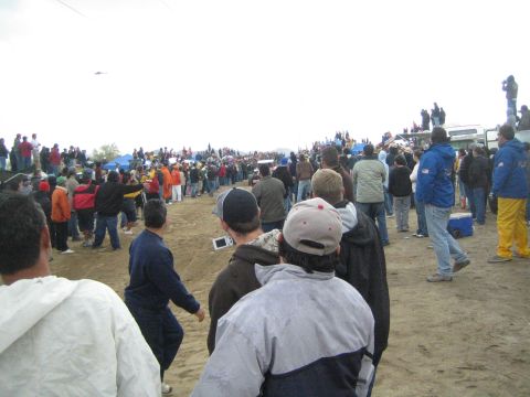 1990 Ford Bronco - Spectators in jackets and caps gather along a dirt course at an off-road desert race under a flat gray sky.