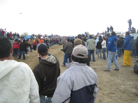 1990 Ford Bronco - line of bundled-up spectators watches an off-road race along a sandy dirt course beneath an overcast sky.