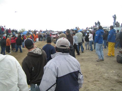 1990 Ford Bronco - red and white off-road race car moves along a distant dirt course as spectators line both sides under gray skies.