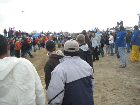 1990 Ford Bronco - Spectators in jackets and caps line a dirt off-road course, watching a race car pass in the distance under an overcast sky.