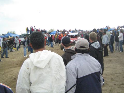 1990 Ford Bronco - crowd of spectators lines a dirt course as a small off-road race car passes during a desert race event.