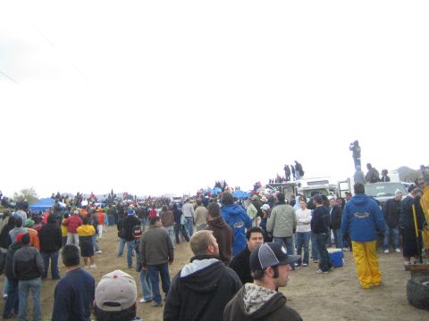 1990 Ford Bronco - sprawling crowd of spectators fills a dirt off-road course, with tents, trucks and people on rooftops watching under gray skies.