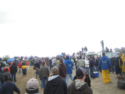 1990 Ford Bronco - Spectators spread across a wide dirt off-road course, some standing on trucks, watching the race under an overcast sky.