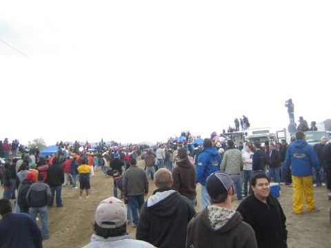 1990 Ford Bronco - broad crowd of spectators in jackets and caps gathers along a sandy off-road course at a desert race.