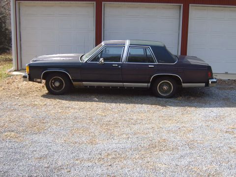1982 Ford LTD Crown Victoria - Dark blue full-size Ford four-door sedan shot in full profile on gravel in front of a red barn with three white roll-up garage doors. It ha…