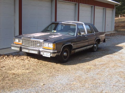1982 Ford LTD Crown Victoria - Front three-quarter view of a weathered gray-blue Ford LTD Crown Victoria four-door sedan parked on gravel in front of a white-and-red pole…