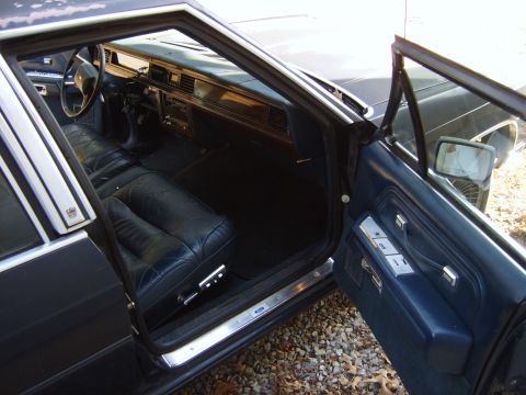 1982 Ford LTD Crown Victoria - Driver's door open on a blue-interior Ford sedan, looking down at a worn dark blue leather split bench seat, blue carpet, and a woodgrain-t…