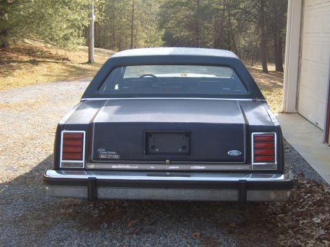 1982 Ford LTD Crown Victoria - Straight-on rear view of a dark blue Ford LTD Crown Victoria sedan with a padded vinyl half-roof, parked on gravel beside a garage with pin…
