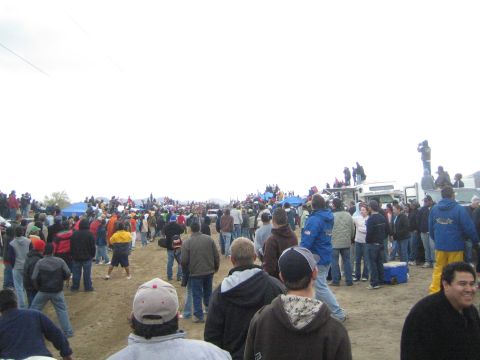 1990 Ford Bronco - Spectators line a dirt off-road course dotted with blue tents and trucks, watching a race under an overcast sky.
