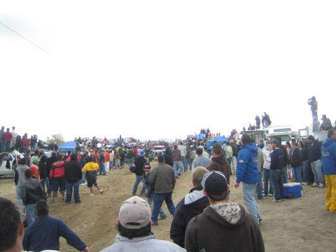 1990 Ford Bronco - large crowd of bundled spectators gathers along a dirt off-road course, with trucks and tents visible in the distance.