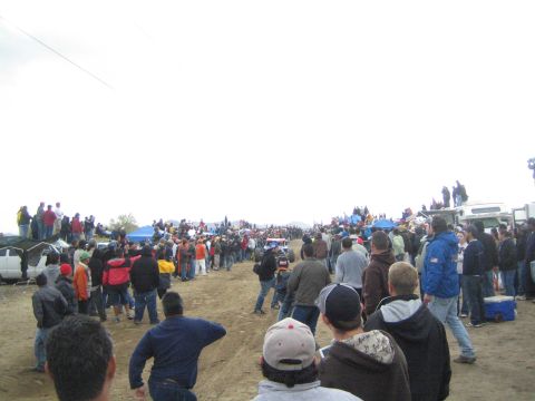 1990 Ford Bronco - Spectators in cold-weather gear line a wide sandy off-road course, watching the race beneath a flat overcast sky.