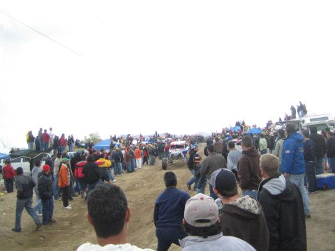 1990 Ford Bronco - small off-road buggy drives along a sandy course lined by a dense crowd of spectators at a desert race.