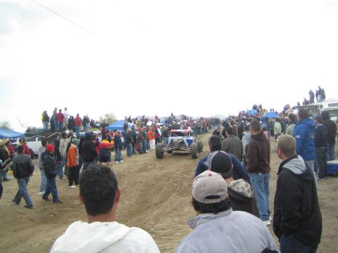 1990 Ford Bronco - yellow single-seat off-road buggy drives down a dirt course as crowds of spectators look on under an overcast sky.