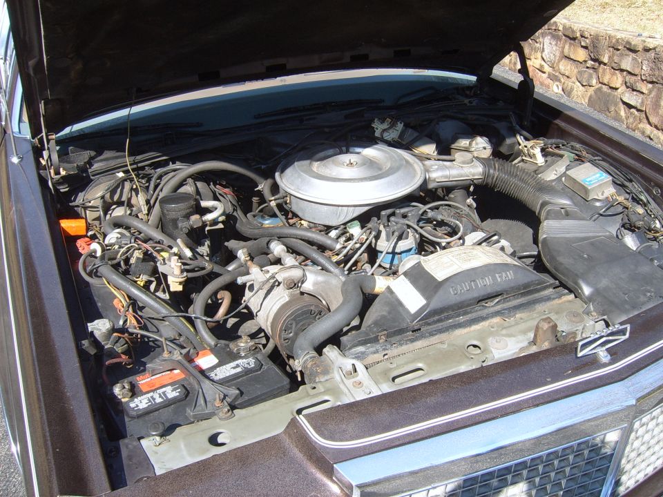 Brown 1980s sedan engine bay with V8 engine, air cleaner, battery, and hoses visible