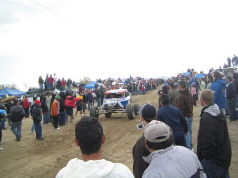 1990 Ford Bronco - blue and white open-wheel off-road buggy heads up a sandy course lined on both sides by spectators at a desert race.