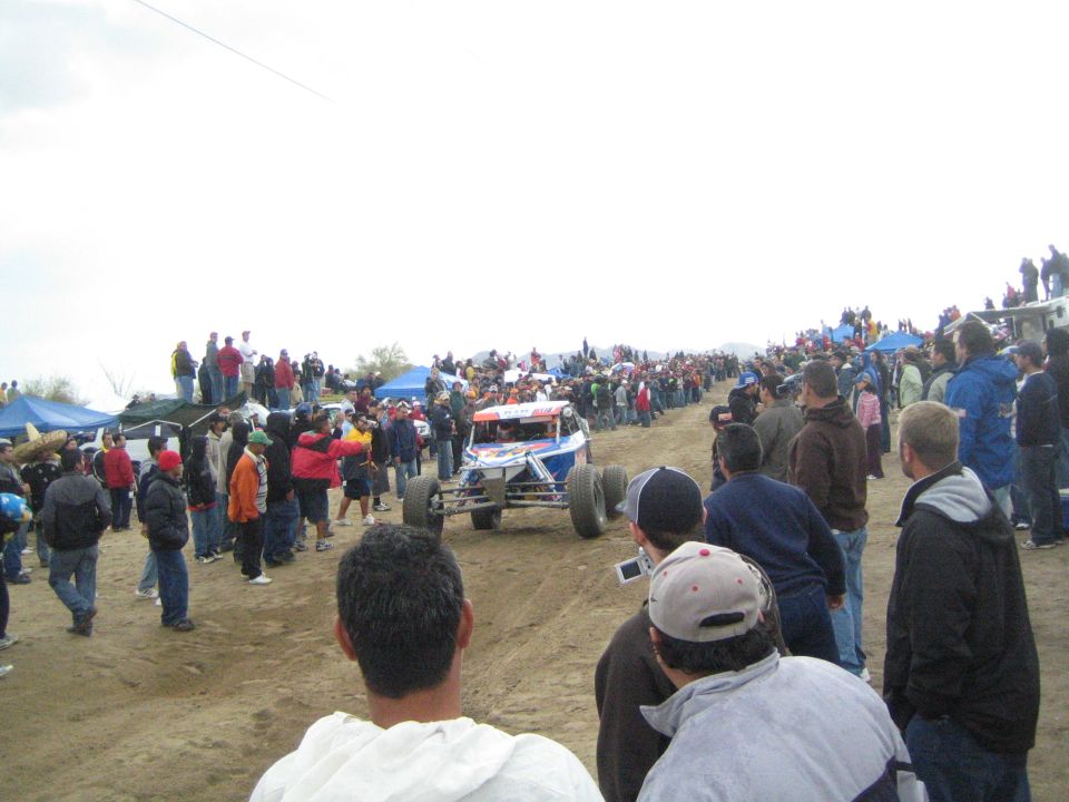 Blue and white off-road race buggy driving up dirt course lined with crowds of spectators