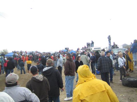 1990 Ford Bronco - large crowd of bundled-up spectators stands on packed dirt near tents and trucks at an overcast outdoor desert event.