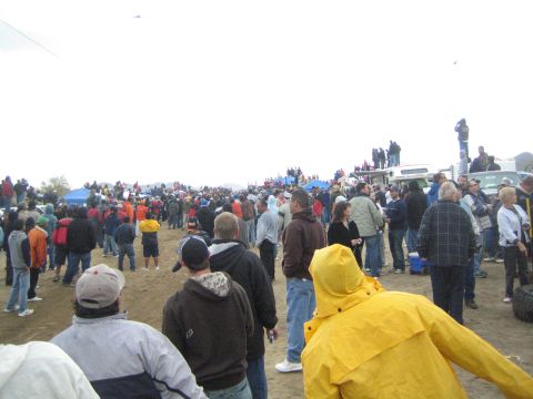 1990 Ford Bronco - Spectators gather on dirt under a gray sky, including a person in a bright yellow rain jacket, near tents and parked vehicles.