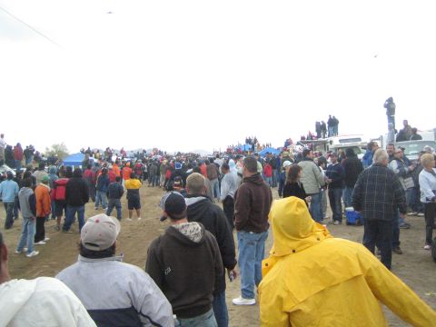 1990 Ford Bronco - dense crowd of spectators stands across an open dirt area dotted with blue tents and parked trucks under an overcast sky.