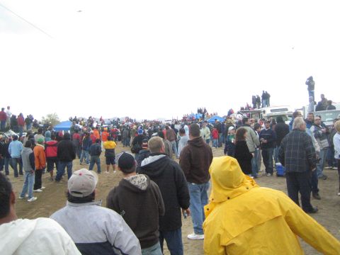 1990 Ford Bronco - Spectators in cold-weather clothing crowd an open dirt area with blue tents and parked vehicles under a gray overcast sky.