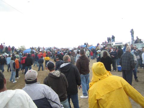 1990 Ford Bronco - large crowd of spectators spreads across packed dirt near tents and trucks, with a person in a yellow rain jacket in the foreground.
