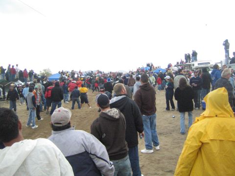 1990 Ford Bronco - Spectators in jackets and caps cluster on an open dirt expanse beside blue tents and parked trucks under an overcast sky.