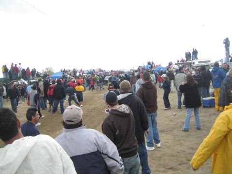 1990 Ford Bronco - wide crowd of spectators is spread across a dirt field with blue tents and parked vehicles beneath a gray sky.