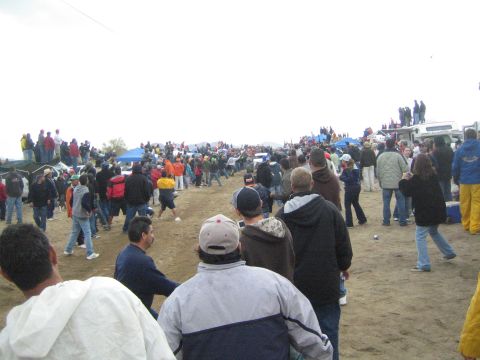 1990 Ford Bronco - Spectators line a wide dirt course dotted with blue tents and parked trucks under an overcast desert sky.