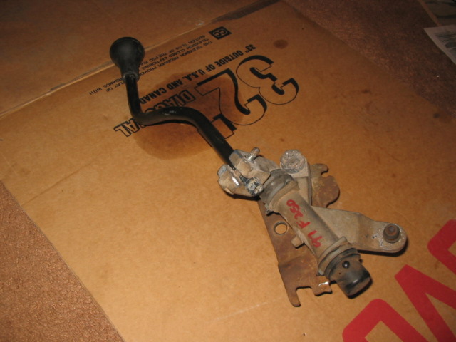 Manual shifter lever and metal mounting base on cardboard, with red marker writing on housing.