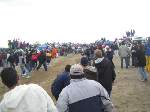 1990 Ford Bronco - Spectators line both sides of a dirt course where a small vehicle moves in the distance, with tents and trucks under a gray sky.