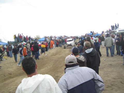 1990 Ford Bronco - white off-road buggy travels along a dirt course lined with crowds of spectators, tents, and trucks under an overcast sky.