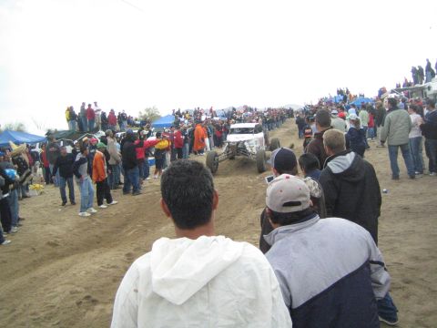 1990 Ford Bronco - white off-road race buggy drives up a dirt course flanked by large crowds of spectators under an overcast desert sky.