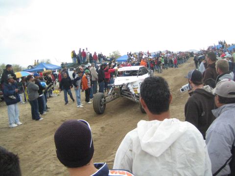 1990 Ford Bronco - white off-road race buggy sits on packed dirt surrounded by spectators, blue tents, and parked vehicles under a gray sky.
