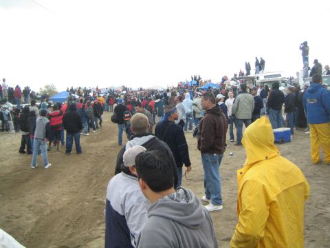 1990 Ford Bronco - Spectators in cold-weather gear stand on open dirt near tents and trucks, with a person in a yellow rain jacket at right, under a gray sky.