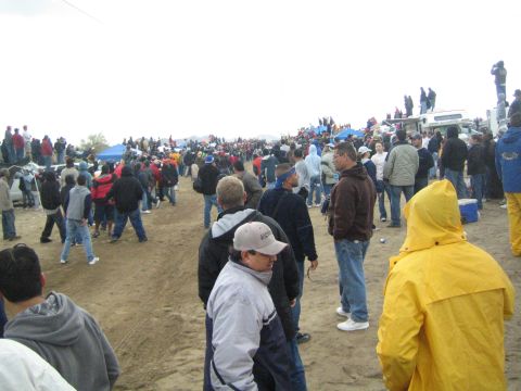 1990 Ford Bronco - crowd of spectators stands on packed dirt near tents and parked trucks, with a person in a yellow rain jacket in the foreground.