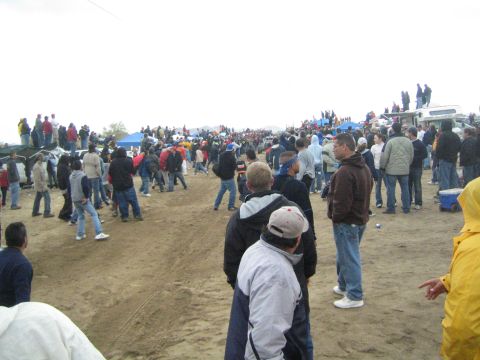 1990 Ford Bronco - Spectators are spread across an open dirt area beside blue tents and parked trucks under an overcast desert sky.