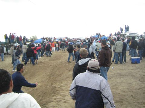 1990 Ford Bronco - crowd of spectators gathers along a wide dirt course with blue tents and parked trucks beneath a gray overcast sky.