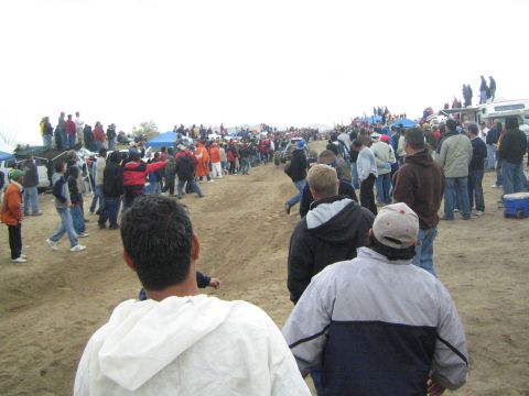 1990 Ford Bronco - Spectators line a wide dirt course dotted with blue tents and parked trucks under an overcast desert sky.