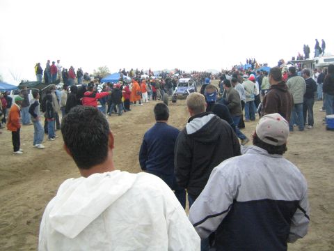 1990 Ford Bronco - Spectators line a dirt course where a small white vehicle moves in the distance, with tents and trucks under an overcast sky.