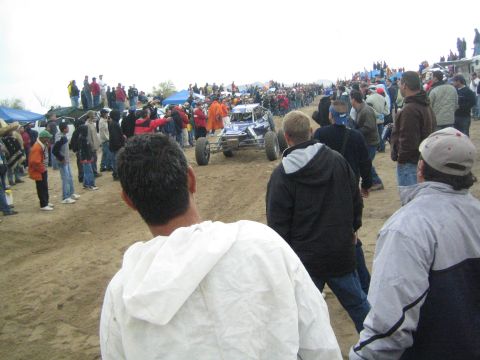 1990 Ford Bronco - white and purple off-road buggy drives along a dirt course flanked by crowds of spectators, tents, and trucks under a gray sky.