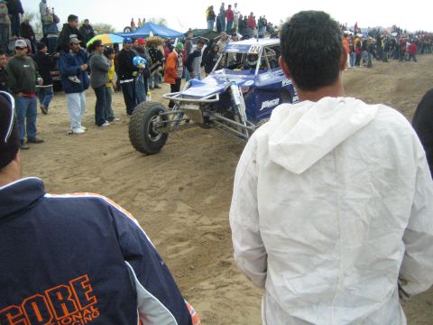 1990 Ford Bronco - blue and white off-road race buggy sits on sandy dirt near spectators and a blue tent, with a person in a SCORE jacket in the foreground.