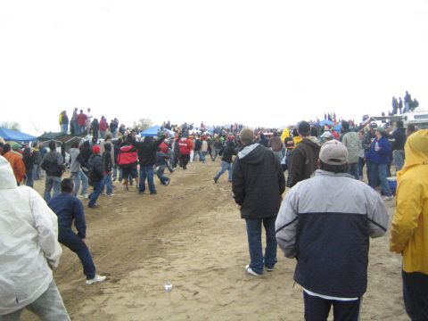 1990 Ford Bronco - crowd of spectators walks along a wide dirt course lined with tents and parked trucks beneath an overcast desert sky.