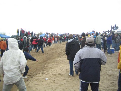 1990 Ford Bronco - Spectators stand across an open dirt course dotted with blue tents and parked trucks under a gray overcast sky.