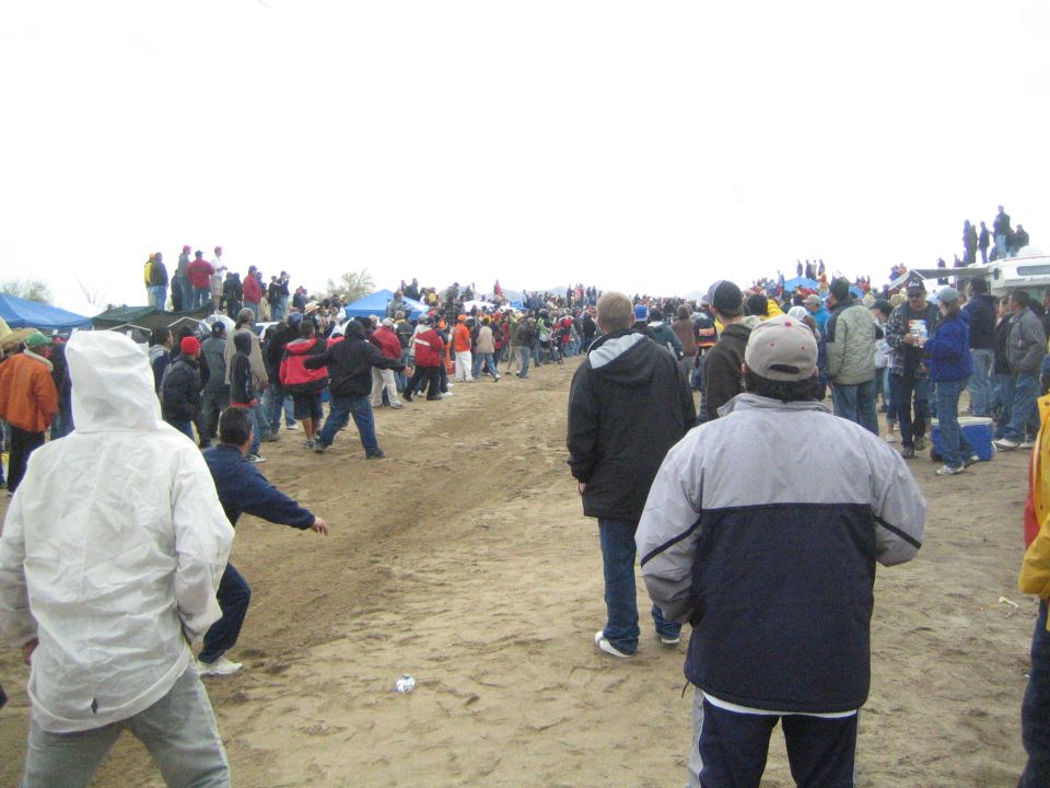 Crowd of spectators on a dirt course with tents and trucks under gray sky