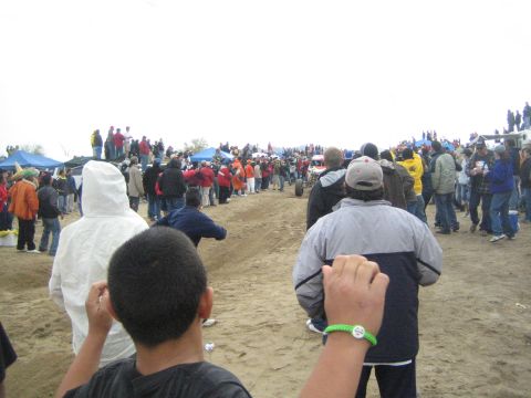 1990 Ford Bronco - Spectators line a dirt course beneath a gray sky, with tents and trucks beyond and a person wearing a green wristband in the foreground.