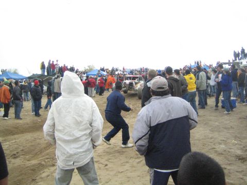 1990 Ford Bronco - Spectators in cold-weather jackets gather on a sandy off-road course as a race buggy passes, with pop-up tents in the background.