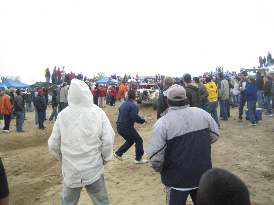 Spectators in jackets watch an off-road race buggy on a sandy course under overcast sky