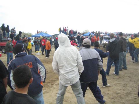 1990 Ford Bronco - crowd in winter jackets stands on sandy ground watching off-road vehicles, with blue pop-up canopies behind them.