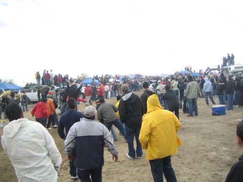 1990 Ford Bronco - Spectators, one in a bright yellow rain jacket, watch an off-road desert race from a sandy berm beside parked trucks and tents.